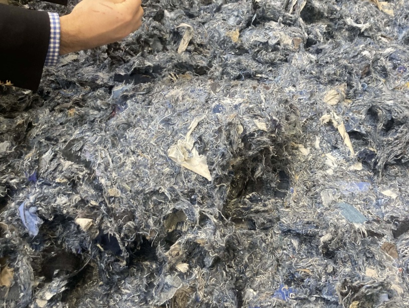 Un amas de fibres textiles recyclées aux teintes majoritairement bleues et grises, composé de résidus de jeans déchiquetés, manipulé par une personne en costume.